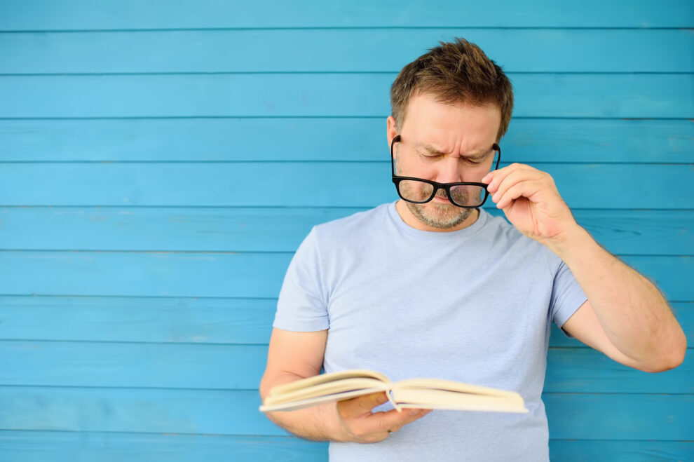 Portrait of mature man with big black eye glasses trying to read book but having difficulties seeing text because of vision problems